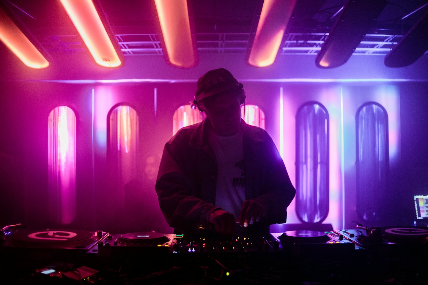 DJ in silhouette behind a mixing board at a neon-lit event.